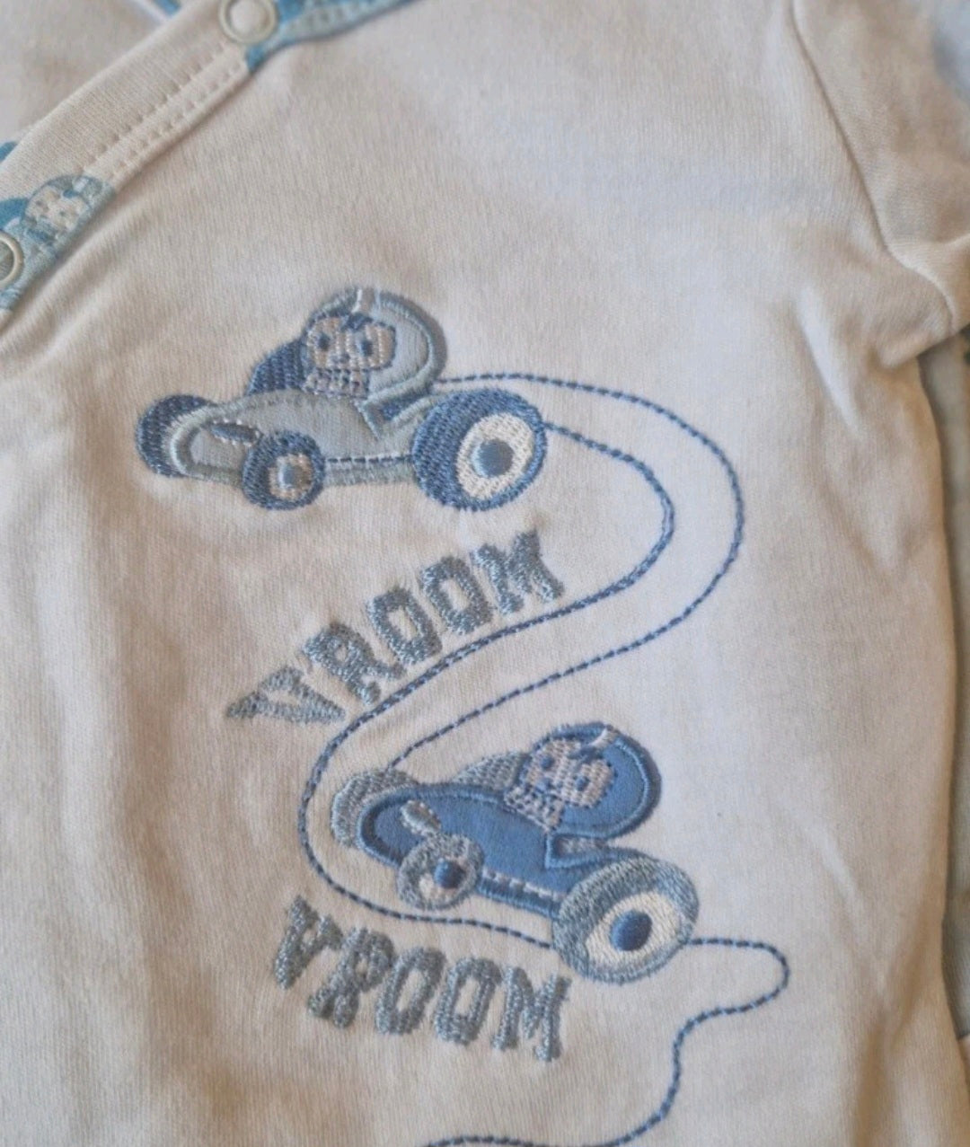 Baby onesie with blue car design and 'Vroom' text on a beige background
