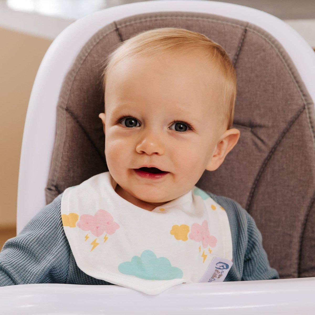 Baby sitting in a high chair wearing a bib with colorful cloud designs.