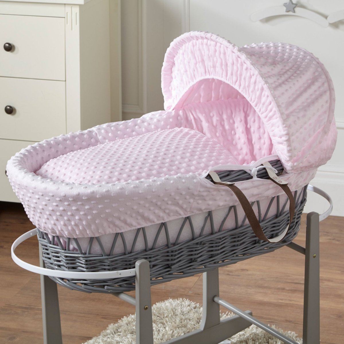 Gray wicker baby Moses basket with pink hood and bedding in a nursery setting.