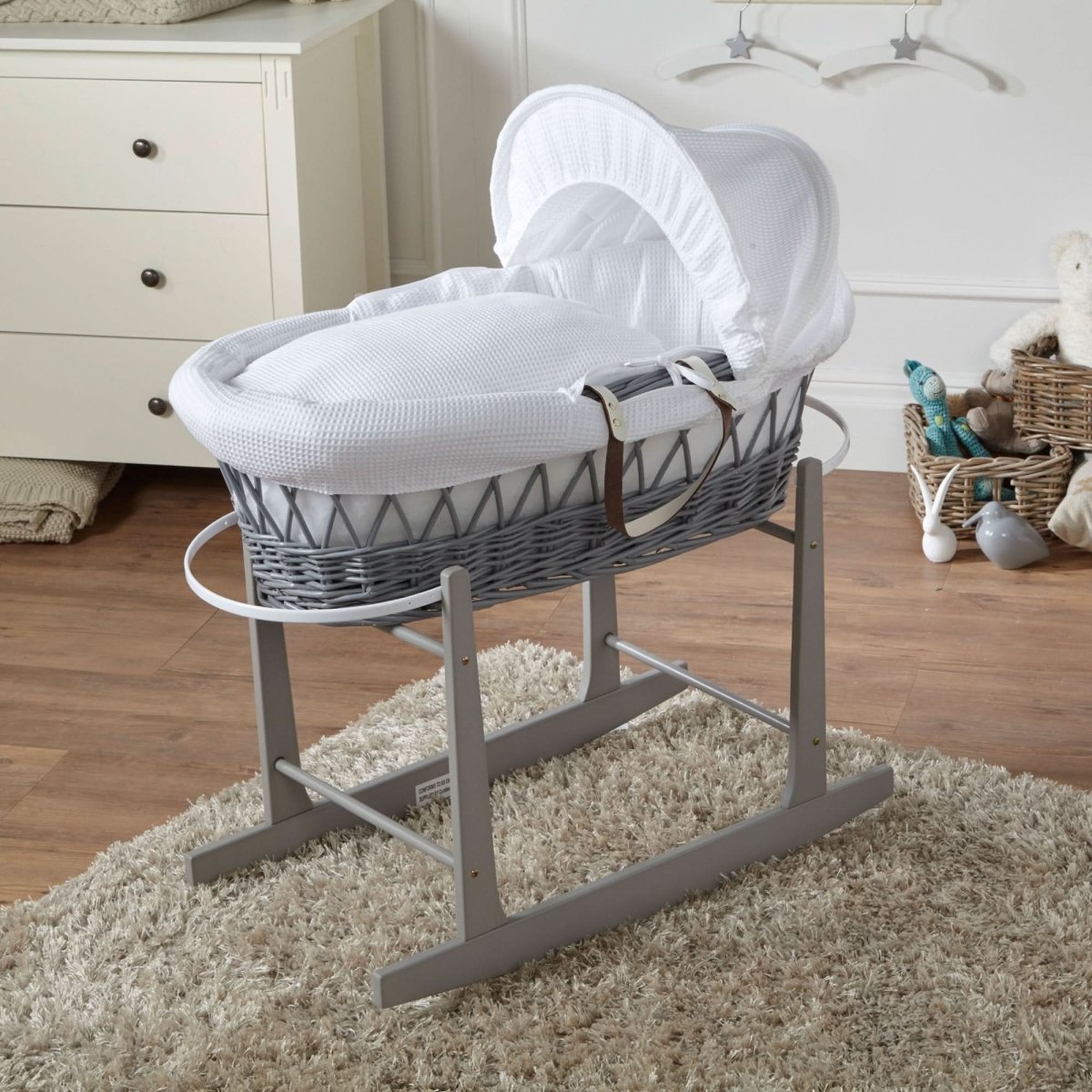 Gray wicker baby Moses basket with white hood on a stand in a nursery setting.